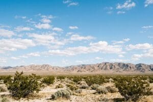 Open Mojave desert scrub below bare mountains and a wide blue sky with scattered white clouds in California's High Desert