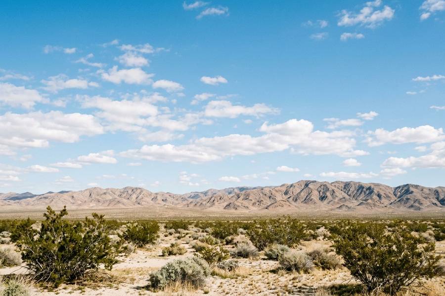 Open Mojave desert scrub below bare mountains and a wide blue sky with scattered white clouds in California's High Desert