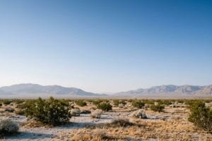 Open Mojave high desert land under a clear blue morning sky, with scattered desert shrubs and bare mountain ranges along the horizon