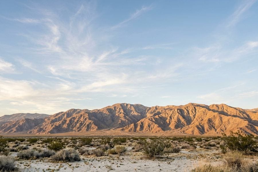 Open desert valley floor in the Coachella Valley with scattered shrubs below sunlit bare mountains and a wide sky with wispy clouds